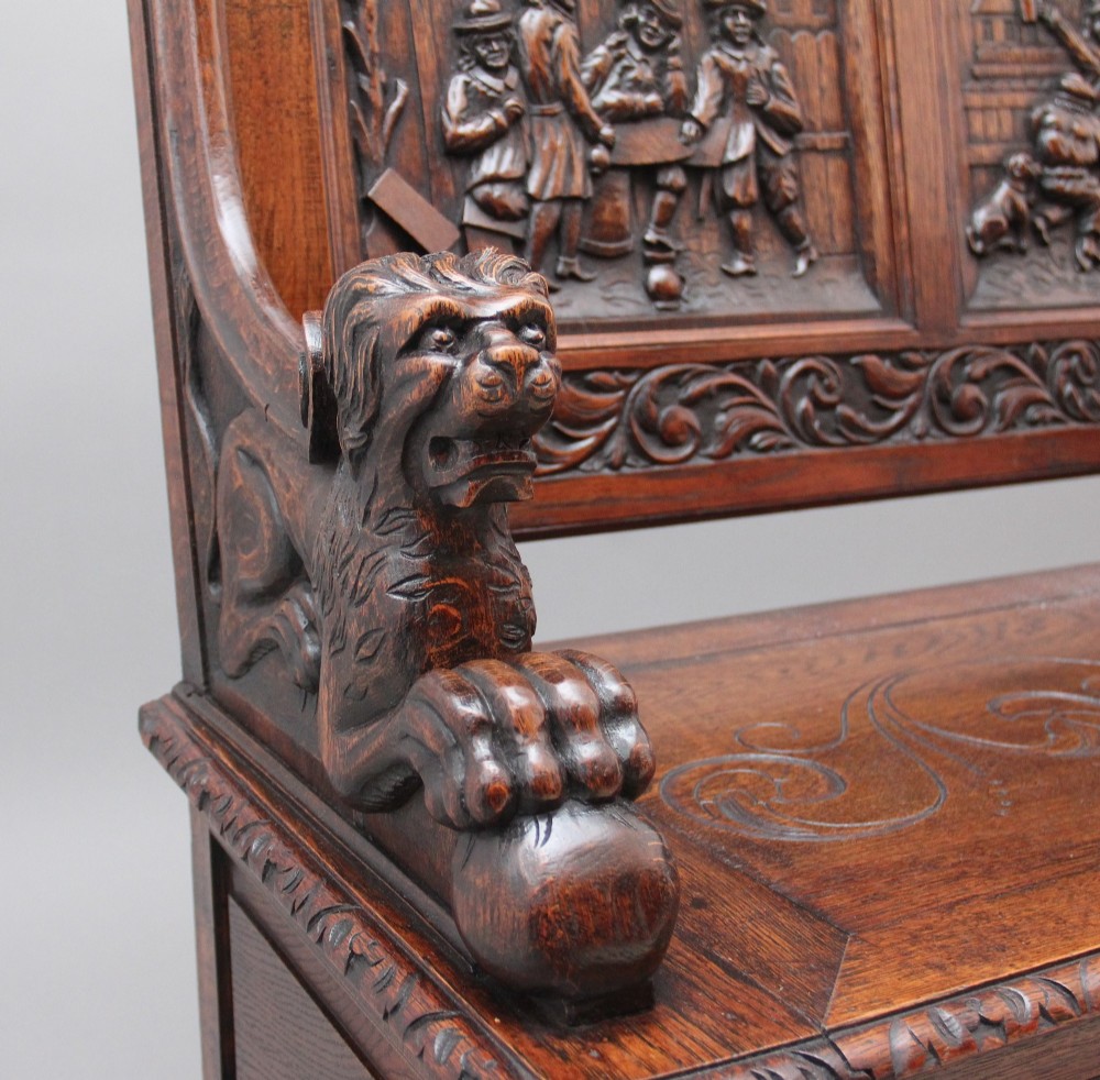 A 19th-century dark oak box settle with intricate relief-carved narrative panels, carved lion head armrests, and a hinged seat for internal storage. View VIII