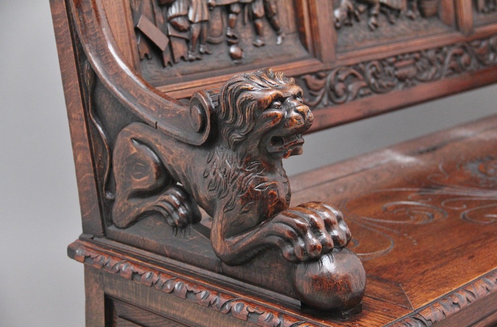 A 19th-century dark oak box settle with intricate relief-carved narrative panels, carved lion head armrests, and a hinged seat for internal storage. View IX