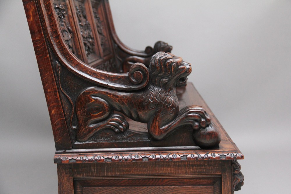 A 19th-century dark oak box settle with intricate relief-carved narrative panels, carved lion head armrests, and a hinged seat for internal storage. View X