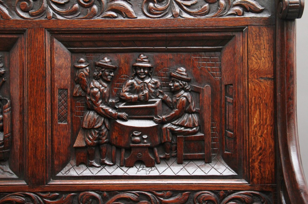 A 19th-century dark oak box settle with intricate relief-carved narrative panels, carved lion head armrests, and a hinged seat for internal storage. View XIV