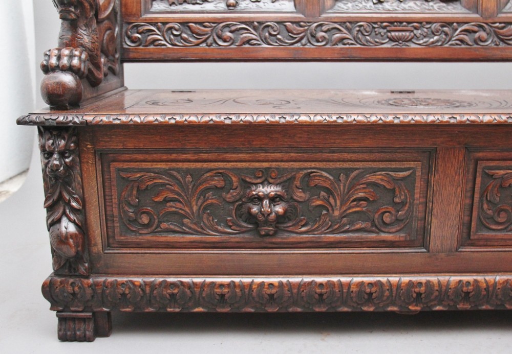 A 19th-century dark oak box settle with intricate relief-carved narrative panels, carved lion head armrests, and a hinged seat for internal storage. View XV