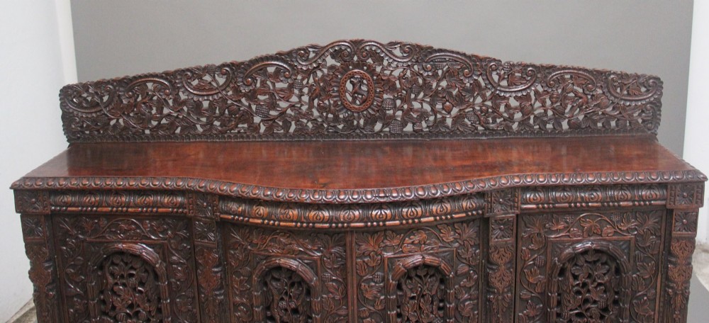 A 19th-century Anglo-Indian dark wood cabinet featuring elaborate pierced foliage carvings, four cupboard doors, and a decorative carved gallery back. View VI
