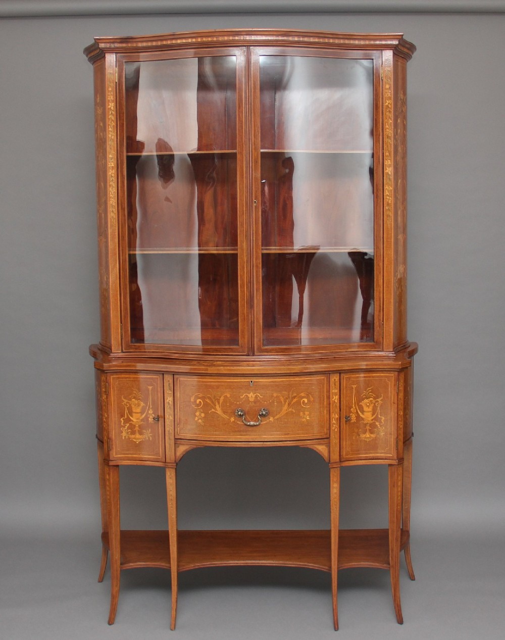 A 19th-century mahogany display cabinet featuring a upper bow-front section with two glazed doors, classical floral satinwood marquetry, a single central drawer, and tapered legs joined by a lower shelf.