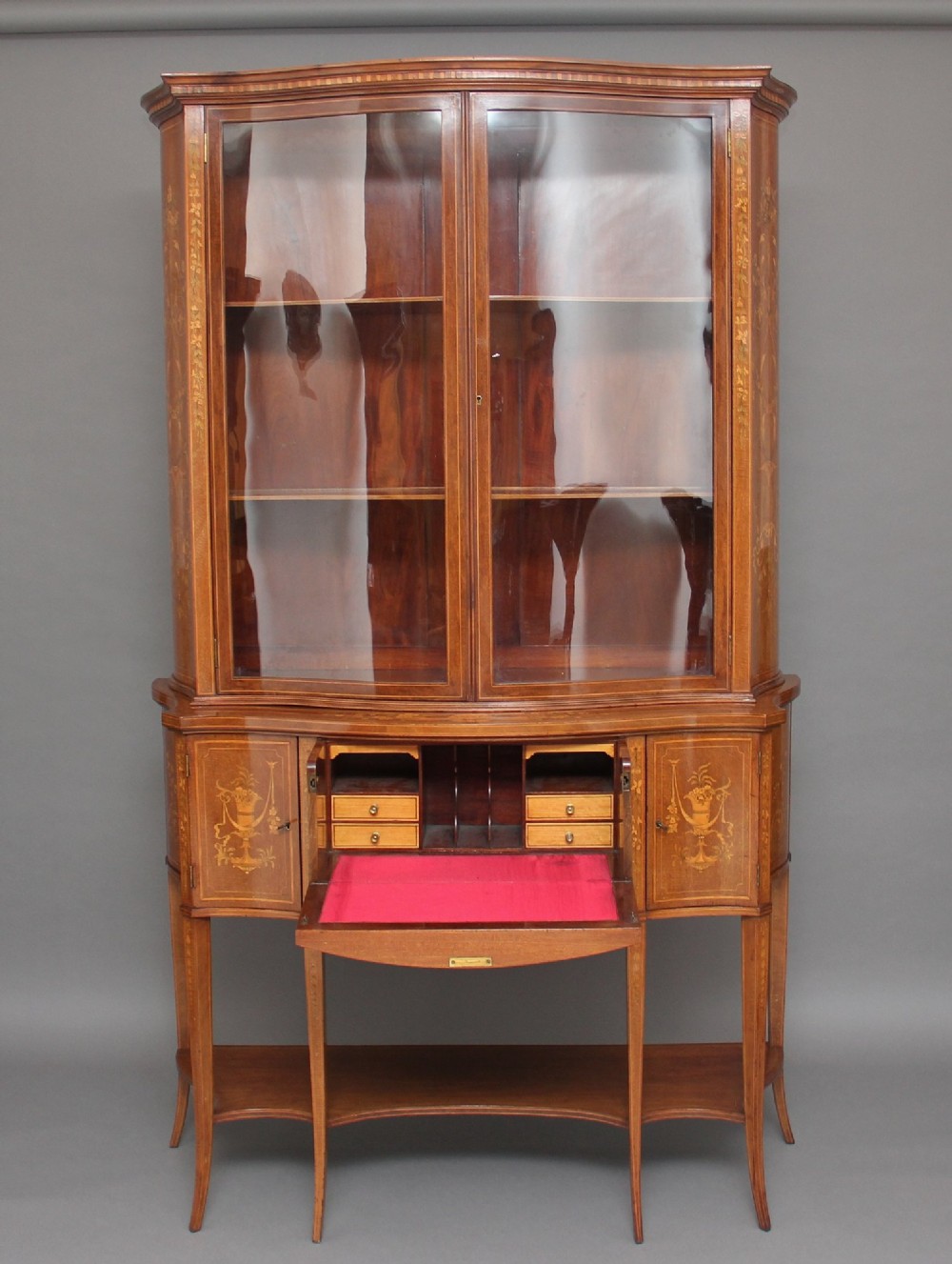 A 19th-century mahogany display cabinet featuring a upper bow-front section with two glazed doors, classical floral satinwood marquetry, a single central drawer, and tapered legs joined by a lower shelf. View II