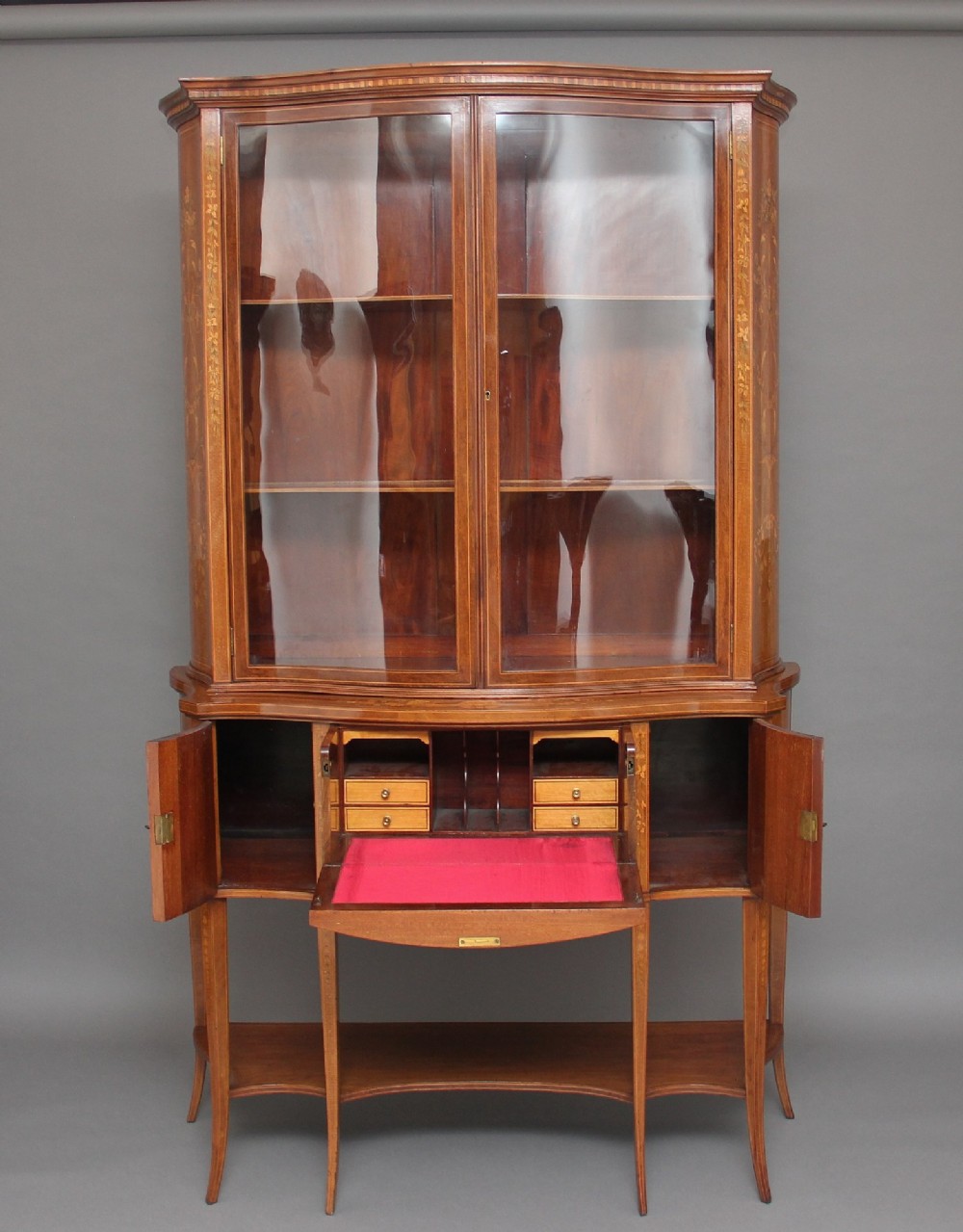 A 19th-century mahogany display cabinet featuring a upper bow-front section with two glazed doors, classical floral satinwood marquetry, a single central drawer, and tapered legs joined by a lower shelf. View III