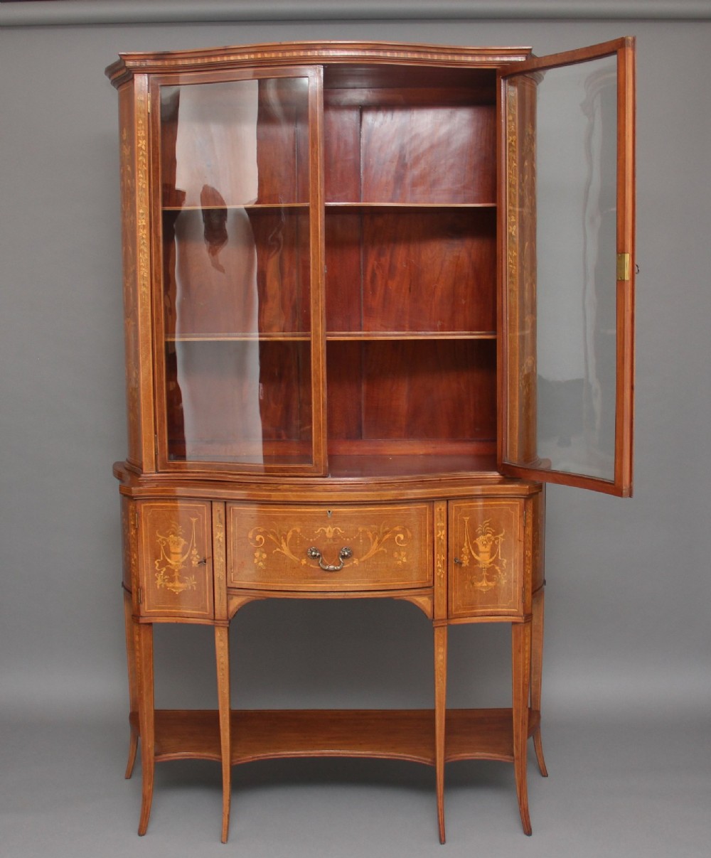 A 19th-century mahogany display cabinet featuring a upper bow-front section with two glazed doors, classical floral satinwood marquetry, a single central drawer, and tapered legs joined by a lower shelf. View IV