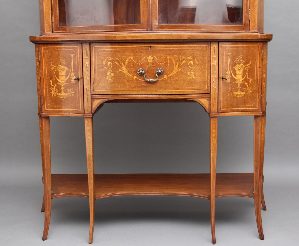 A 19th-century mahogany display cabinet featuring a upper bow-front section with two glazed doors, classical floral satinwood marquetry, a single central drawer, and tapered legs joined by a lower shelf. View V