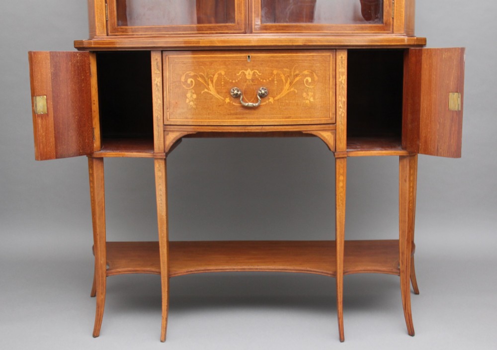 A 19th-century mahogany display cabinet featuring a upper bow-front section with two glazed doors, classical floral satinwood marquetry, a single central drawer, and tapered legs joined by a lower shelf. View VI