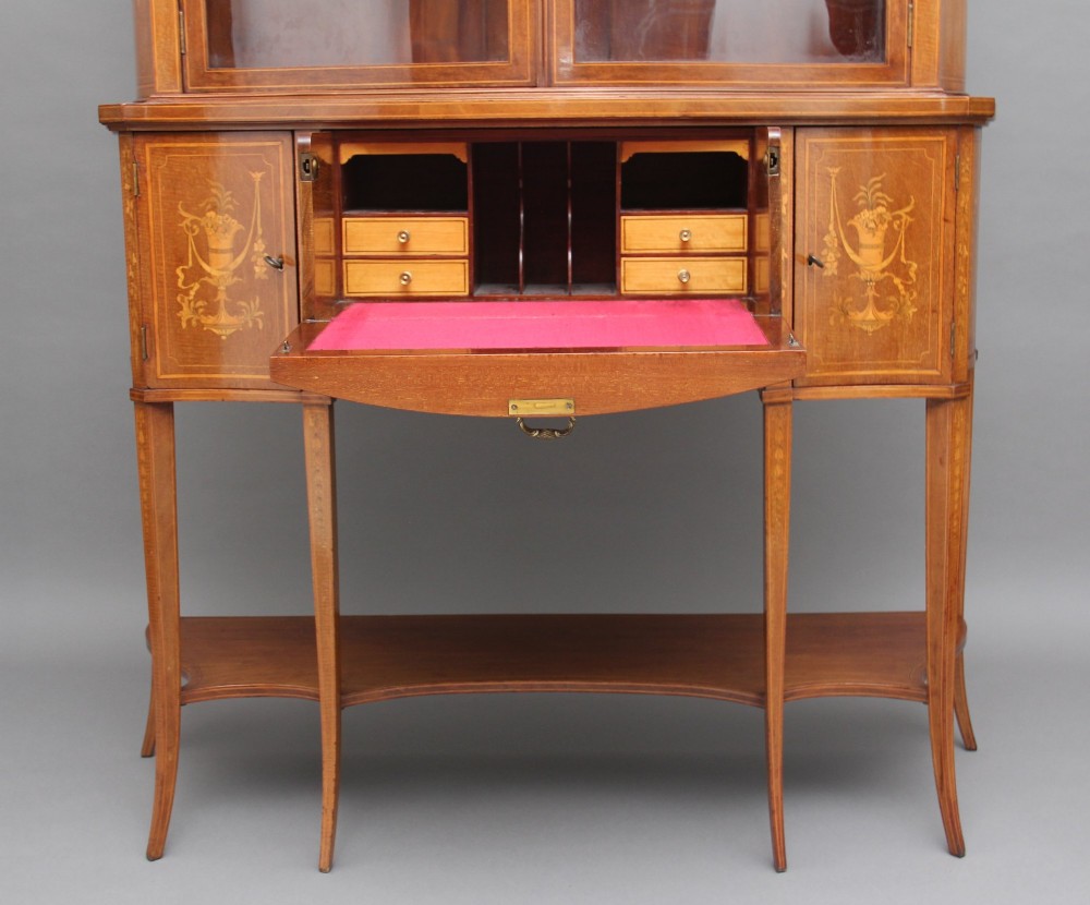 A 19th-century mahogany display cabinet featuring a upper bow-front section with two glazed doors, classical floral satinwood marquetry, a single central drawer, and tapered legs joined by a lower shelf. View VII