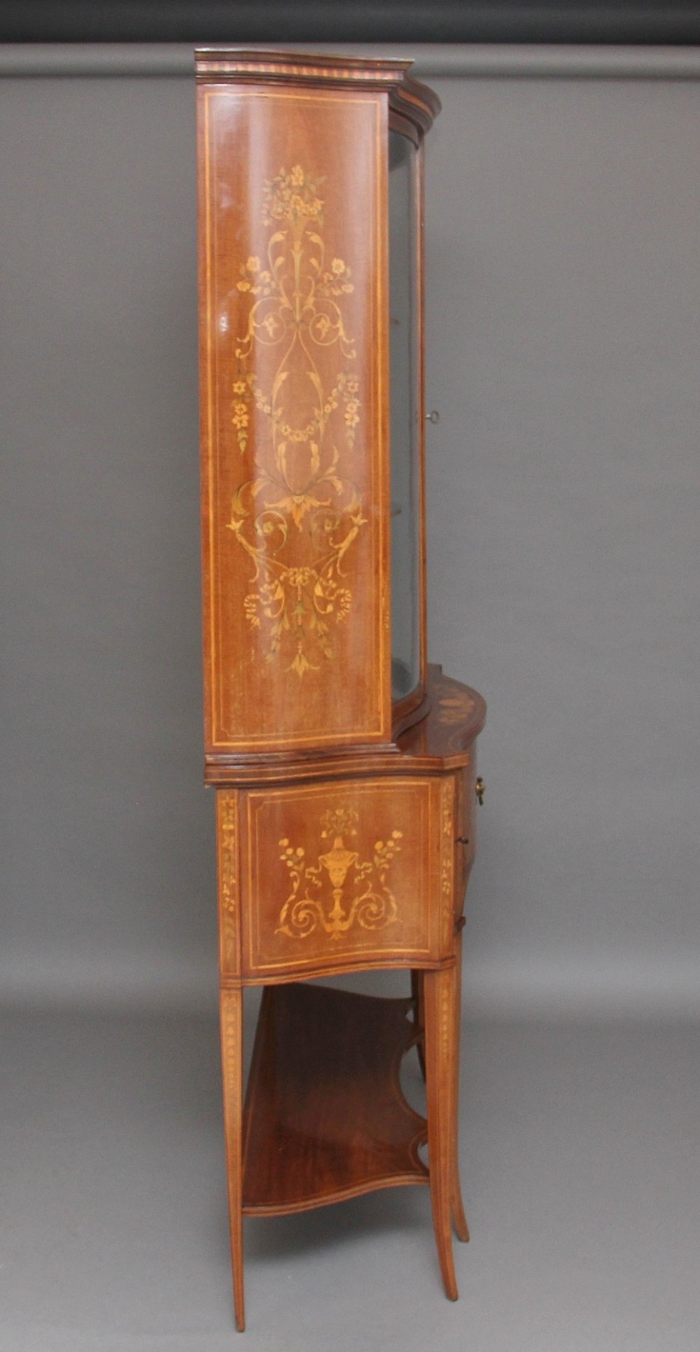 A 19th-century mahogany display cabinet featuring a upper bow-front section with two glazed doors, classical floral satinwood marquetry, a single central drawer, and tapered legs joined by a lower shelf. View IX