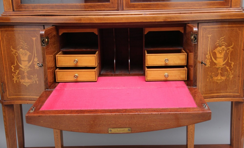 A 19th-century mahogany display cabinet featuring a upper bow-front section with two glazed doors, classical floral satinwood marquetry, a single central drawer, and tapered legs joined by a lower shelf. View X