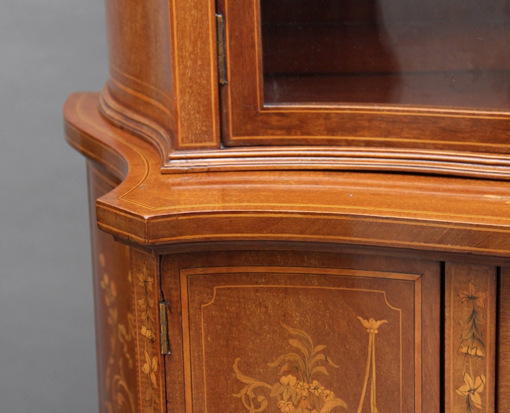 A 19th-century mahogany display cabinet featuring a upper bow-front section with two glazed doors, classical floral satinwood marquetry, a single central drawer, and tapered legs joined by a lower shelf. View XI