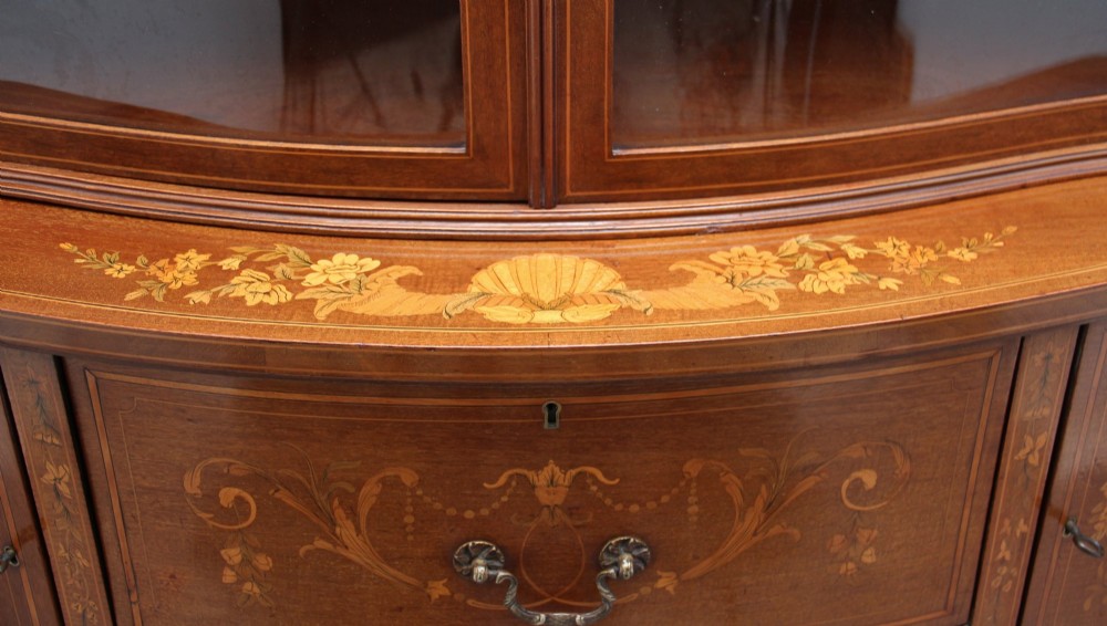 A 19th-century mahogany display cabinet featuring a upper bow-front section with two glazed doors, classical floral satinwood marquetry, a single central drawer, and tapered legs joined by a lower shelf. View XII
