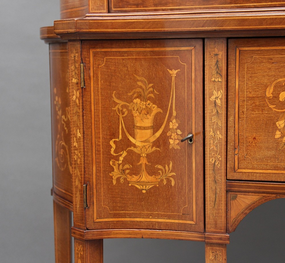A 19th-century mahogany display cabinet featuring a upper bow-front section with two glazed doors, classical floral satinwood marquetry, a single central drawer, and tapered legs joined by a lower shelf. View XIII