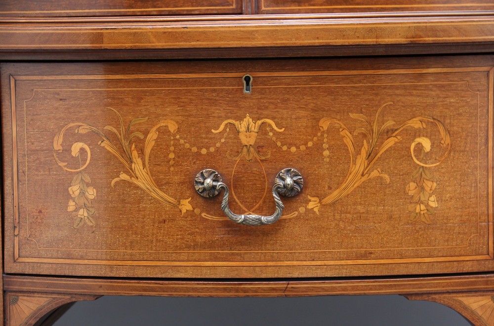 A 19th-century mahogany display cabinet featuring a upper bow-front section with two glazed doors, classical floral satinwood marquetry, a single central drawer, and tapered legs joined by a lower shelf. View XIV