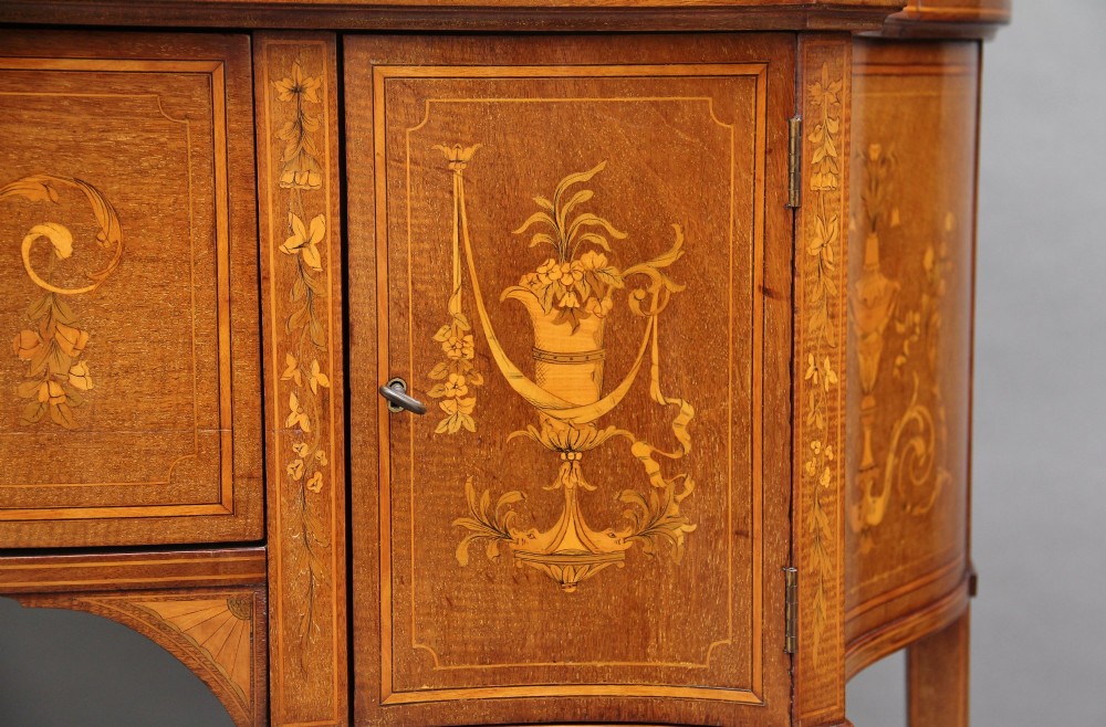 A 19th-century mahogany display cabinet featuring a upper bow-front section with two glazed doors, classical floral satinwood marquetry, a single central drawer, and tapered legs joined by a lower shelf. View XV