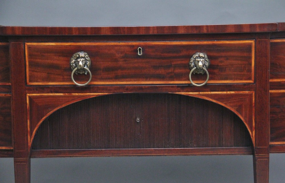 A nineteenth-century mahogany bowfront sideboard featuring inlaid crossbanding, multiple drawers with brass lion-mask ring handles, and tapered square legs. View V
