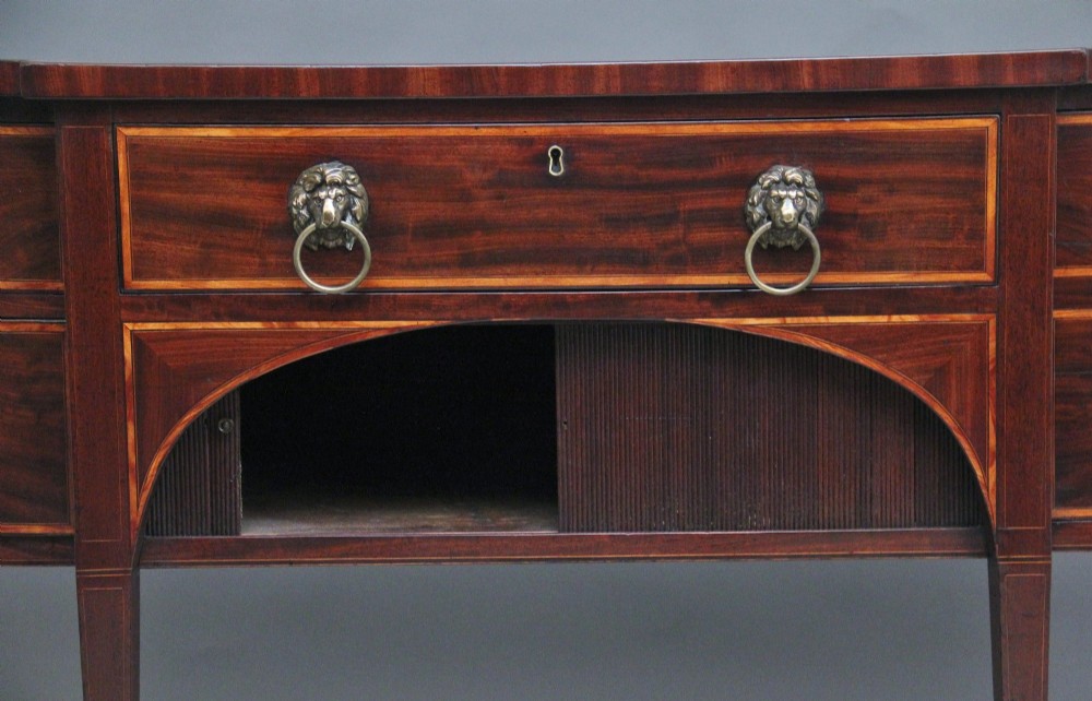 A nineteenth-century mahogany bowfront sideboard featuring inlaid crossbanding, multiple drawers with brass lion-mask ring handles, and tapered square legs. View VI