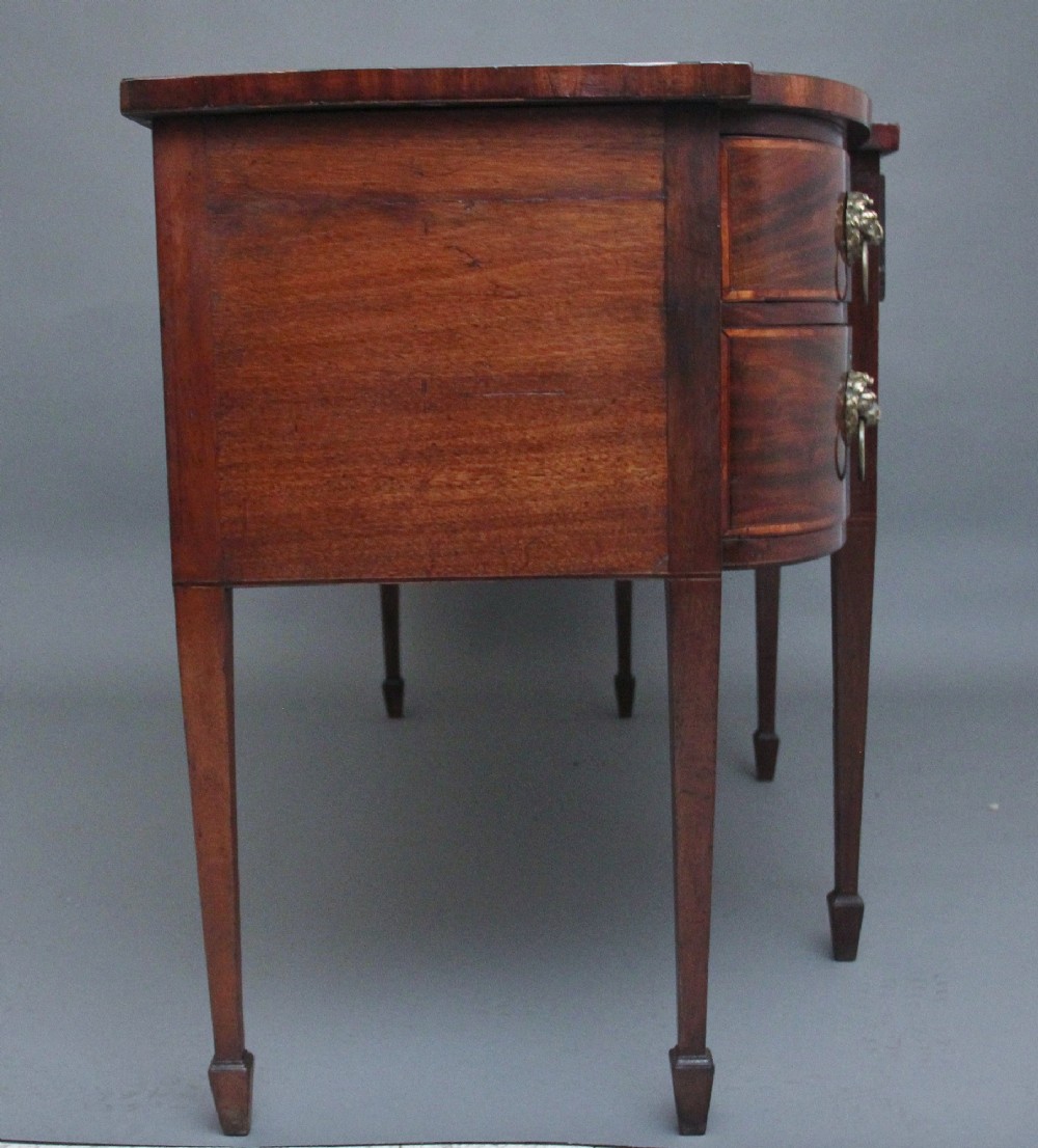 A nineteenth-century mahogany bowfront sideboard featuring inlaid crossbanding, multiple drawers with brass lion-mask ring handles, and tapered square legs. View IX