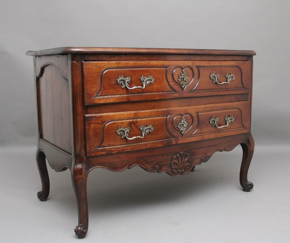 An early nineteenth-century French walnut commode featuring two deep drawers with carved heart-shaped escutcheon surrounds, decorative brass handles, a carved shell apron, and cabriole legs. View II