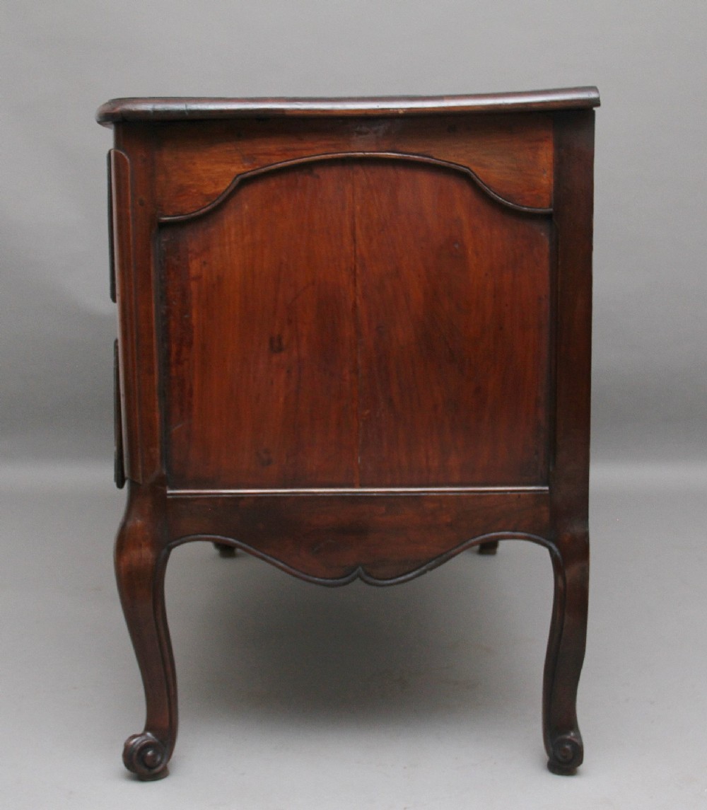 An early nineteenth-century French walnut commode featuring two deep drawers with carved heart-shaped escutcheon surrounds, decorative brass handles, a carved shell apron, and cabriole legs. View VI