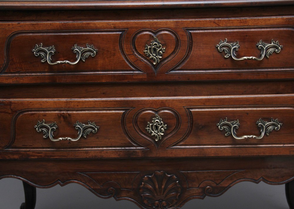 An early nineteenth-century French walnut commode featuring two deep drawers with carved heart-shaped escutcheon surrounds, decorative brass handles, a carved shell apron, and cabriole legs. View IX