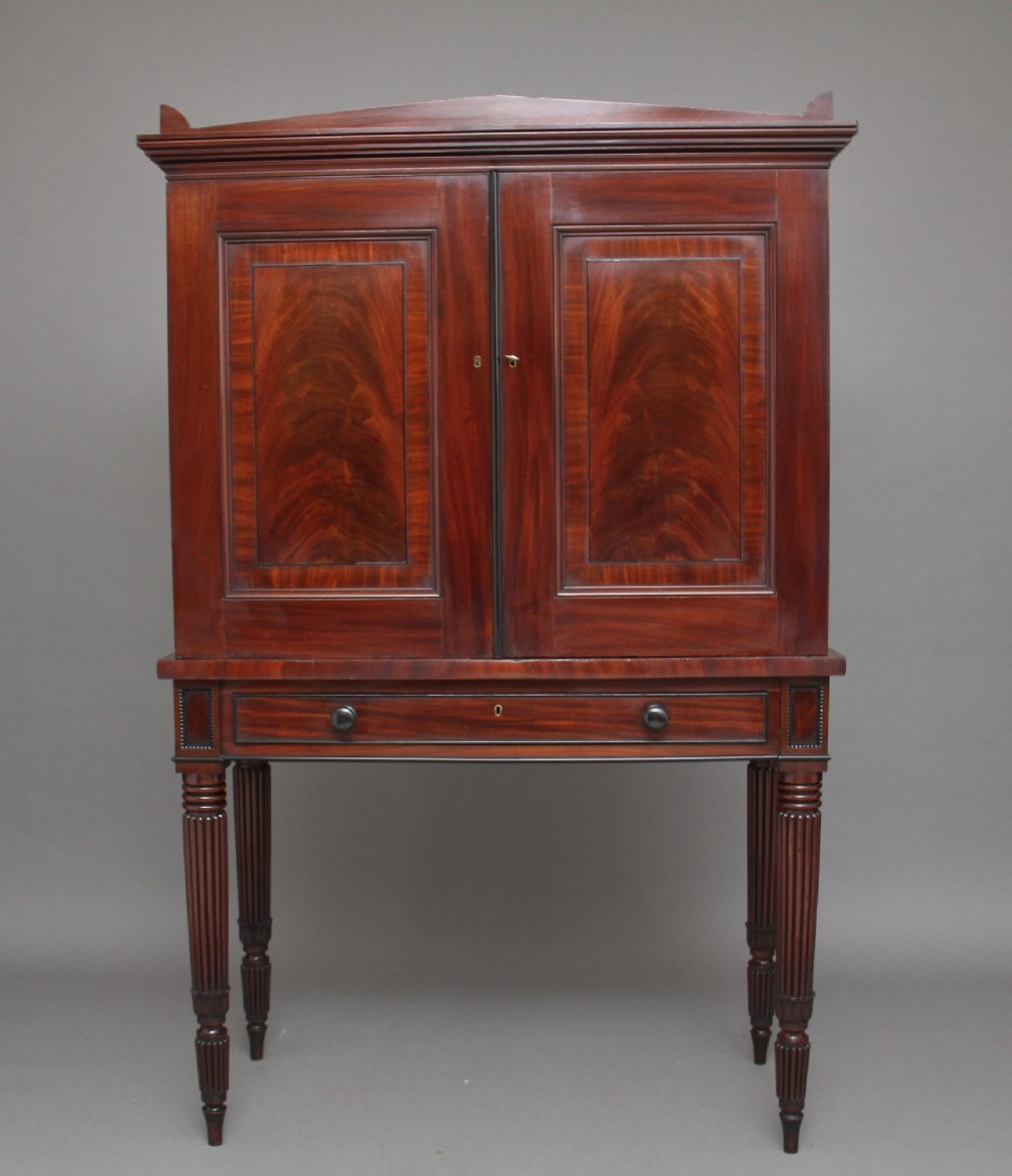 An early nineteenth-century mahogany cabinet on stand featuring flame-veneered panelled double doors above a drawer, supported by turned reeded legs.