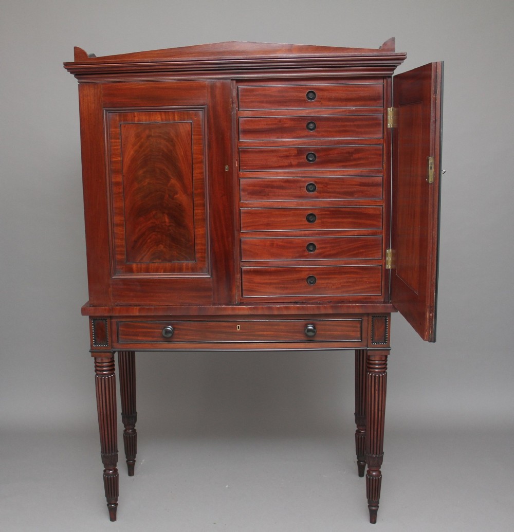 An early nineteenth-century mahogany cabinet on stand featuring flame-veneered panelled double doors above a drawer, supported by turned reeded legs. View II