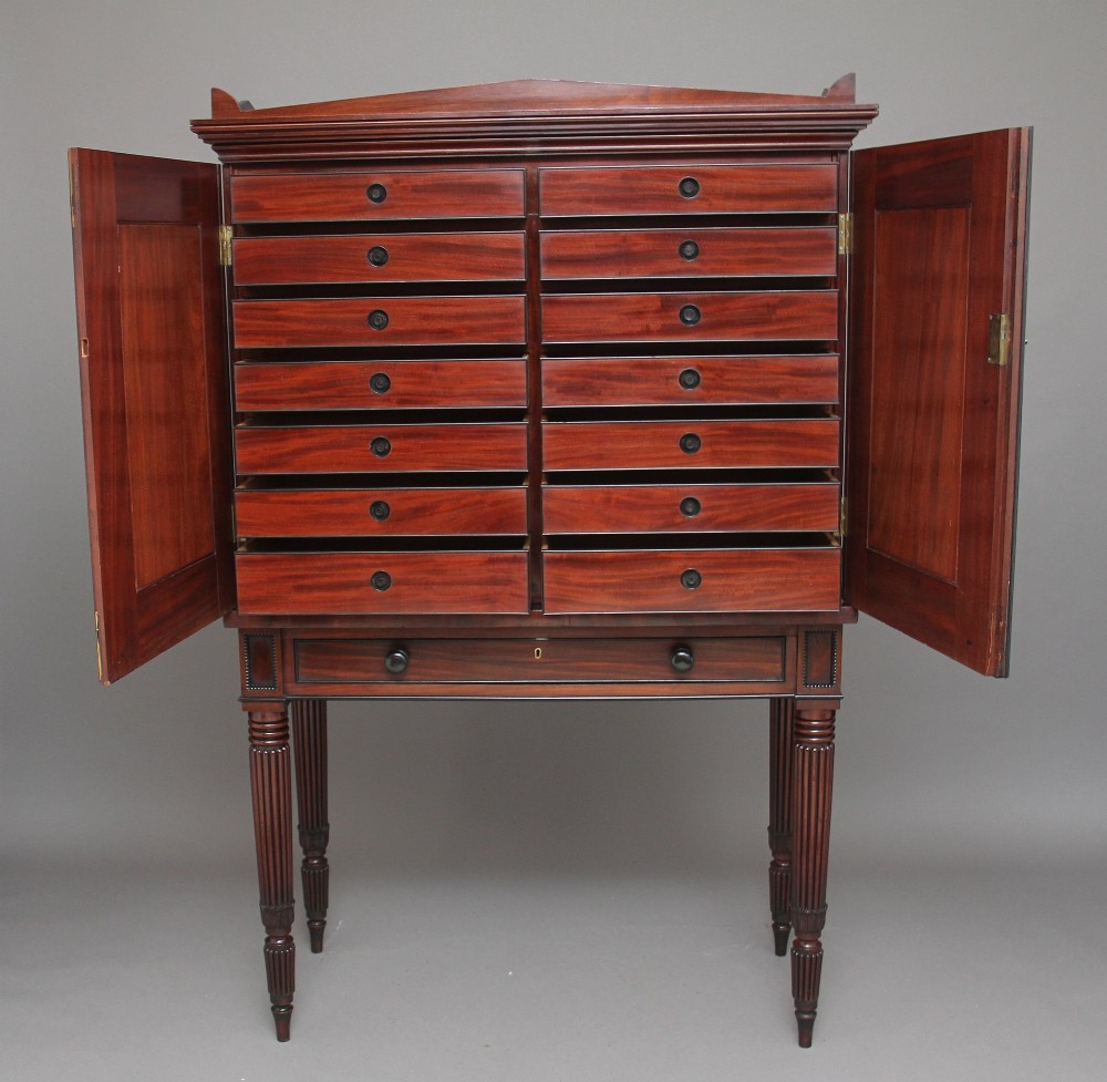 An early nineteenth-century mahogany cabinet on stand featuring flame-veneered panelled double doors above a drawer, supported by turned reeded legs. View VI