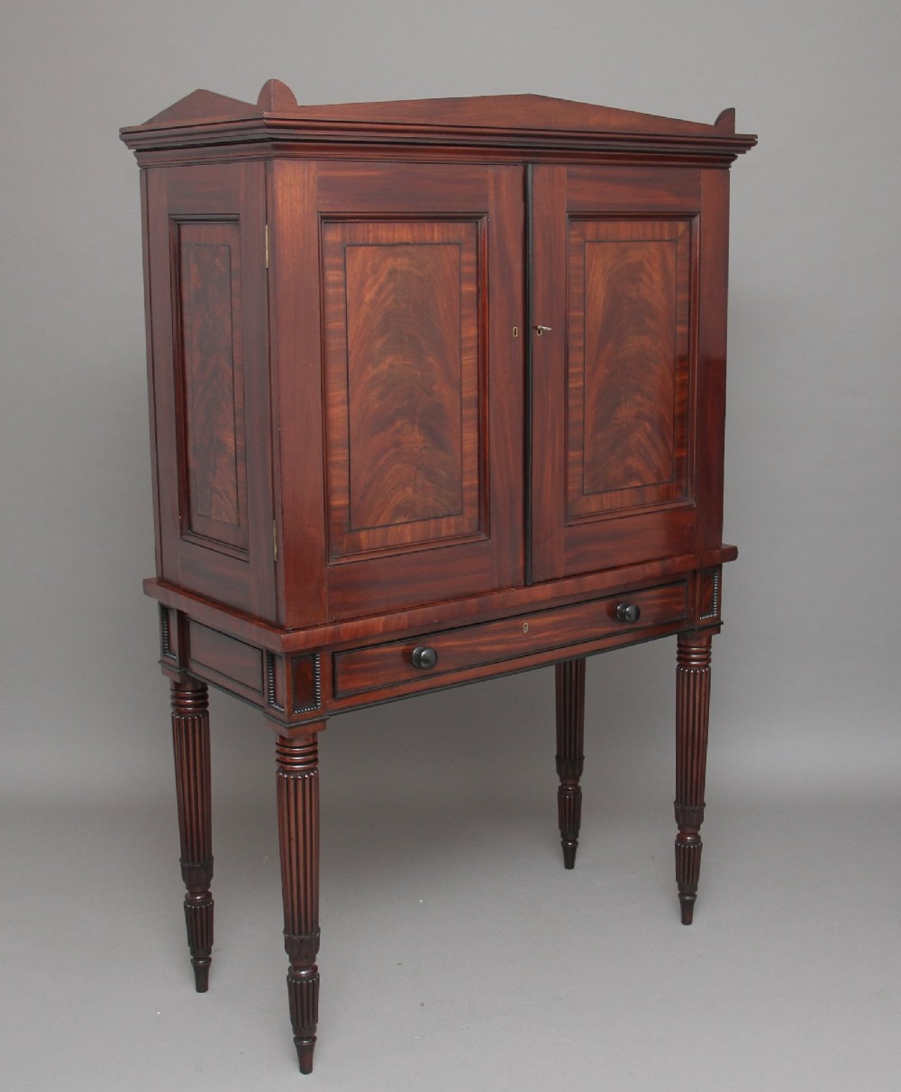 An early nineteenth-century mahogany cabinet on stand featuring flame-veneered panelled double doors above a drawer, supported by turned reeded legs. View VIII