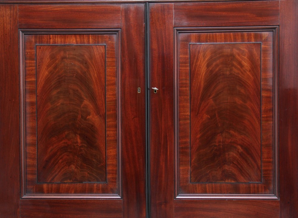 An early nineteenth-century mahogany cabinet on stand featuring flame-veneered panelled double doors above a drawer, supported by turned reeded legs. View IX