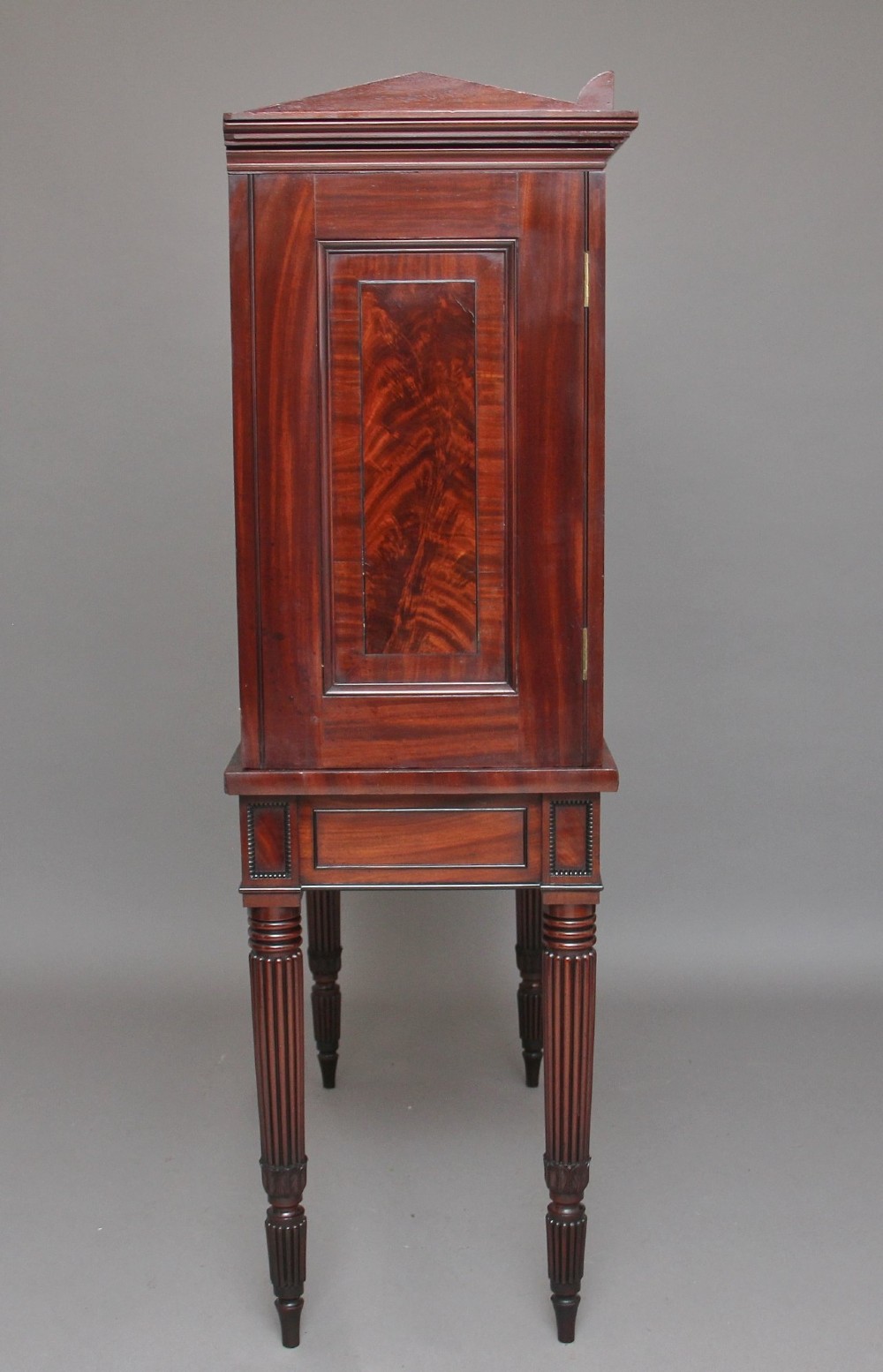 An early nineteenth-century mahogany cabinet on stand featuring flame-veneered panelled double doors above a drawer, supported by turned reeded legs. View X