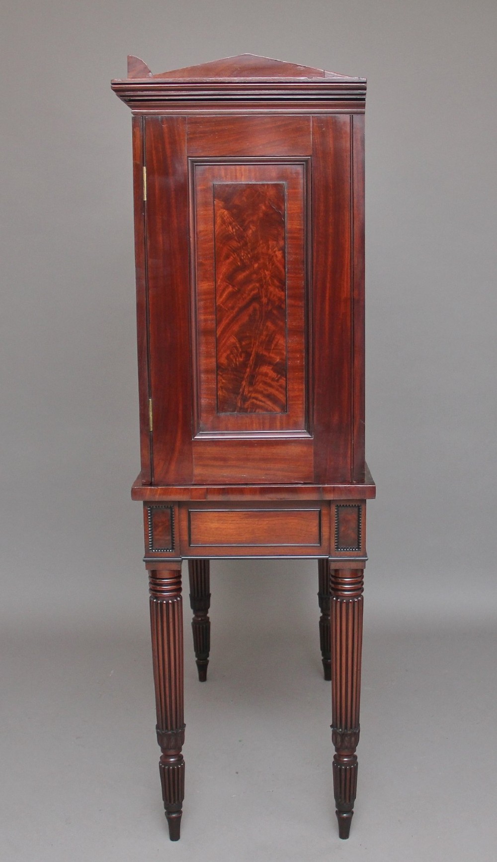 An early nineteenth-century mahogany cabinet on stand featuring flame-veneered panelled double doors above a drawer, supported by turned reeded legs. View XIV