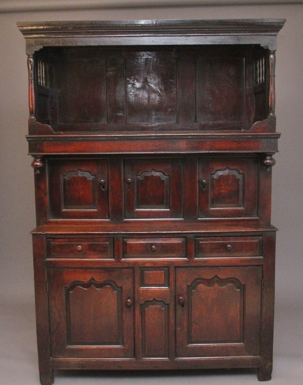 An early eighteenth-century dark Welsh oak tridarn featuring a three-tier design with an open top canopy, middle cupboards with arched panels, and a lower section with drawers and doors.