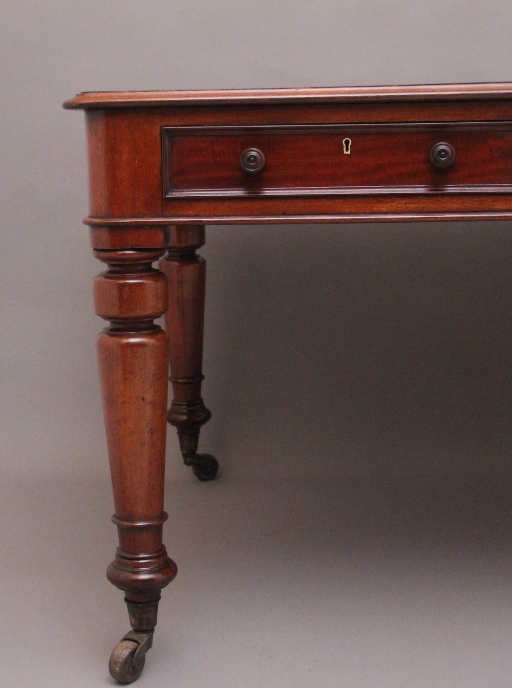 A 19th-century mahogany partners writing table featuring three drawers with turned wooden knobs, an inset leather surface, and four turned legs on brass castors. View IV