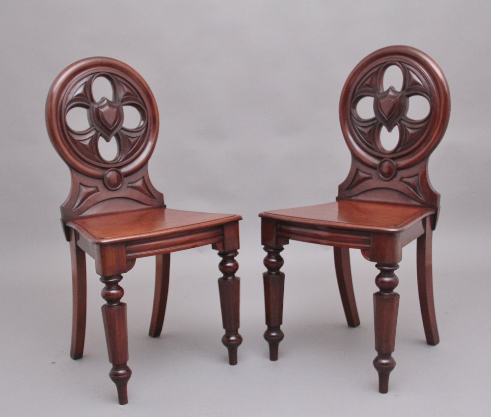 A pair of 19th-century mahogany hall chairs featuring circular carved backrests with Gothic tracery and central shield motifs above solid wooden seats and turned front legs.