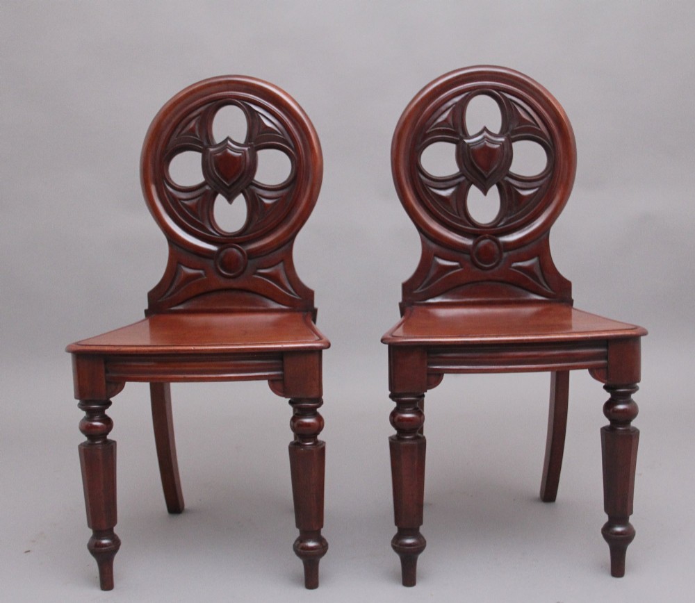 A pair of 19th-century mahogany hall chairs featuring circular carved backrests with Gothic tracery and central shield motifs above solid wooden seats and turned front legs. View II