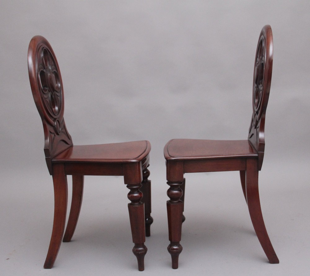 A pair of 19th-century mahogany hall chairs featuring circular carved backrests with Gothic tracery and central shield motifs above solid wooden seats and turned front legs. View III
