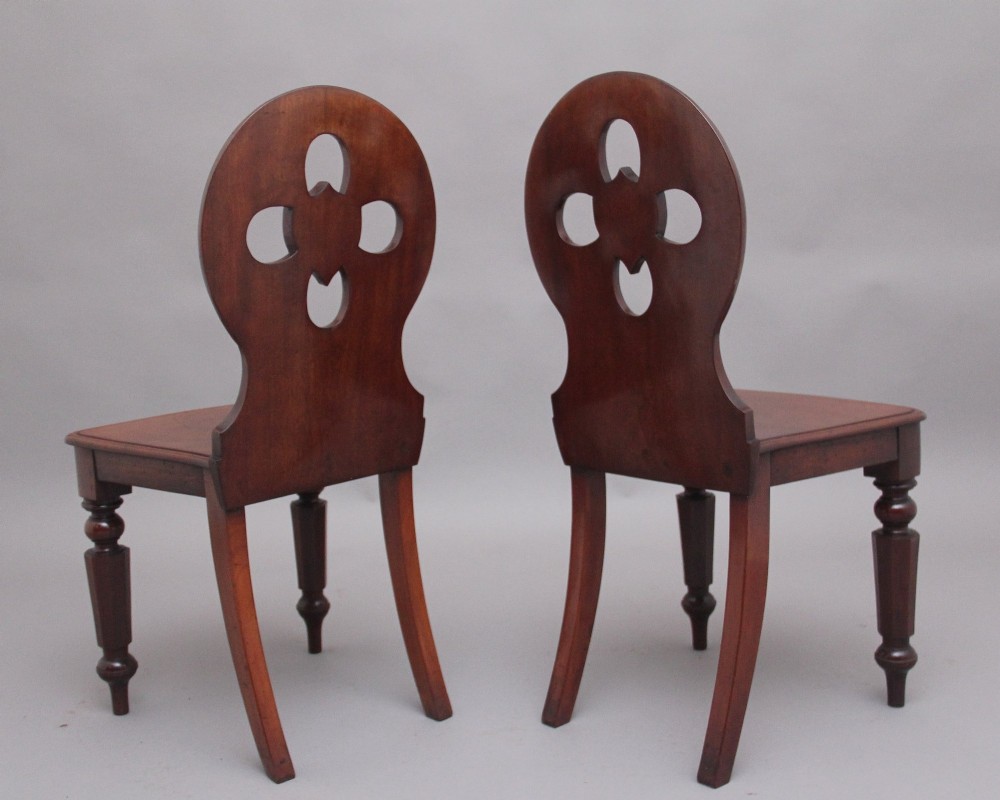 A pair of 19th-century mahogany hall chairs featuring circular carved backrests with Gothic tracery and central shield motifs above solid wooden seats and turned front legs. View IV