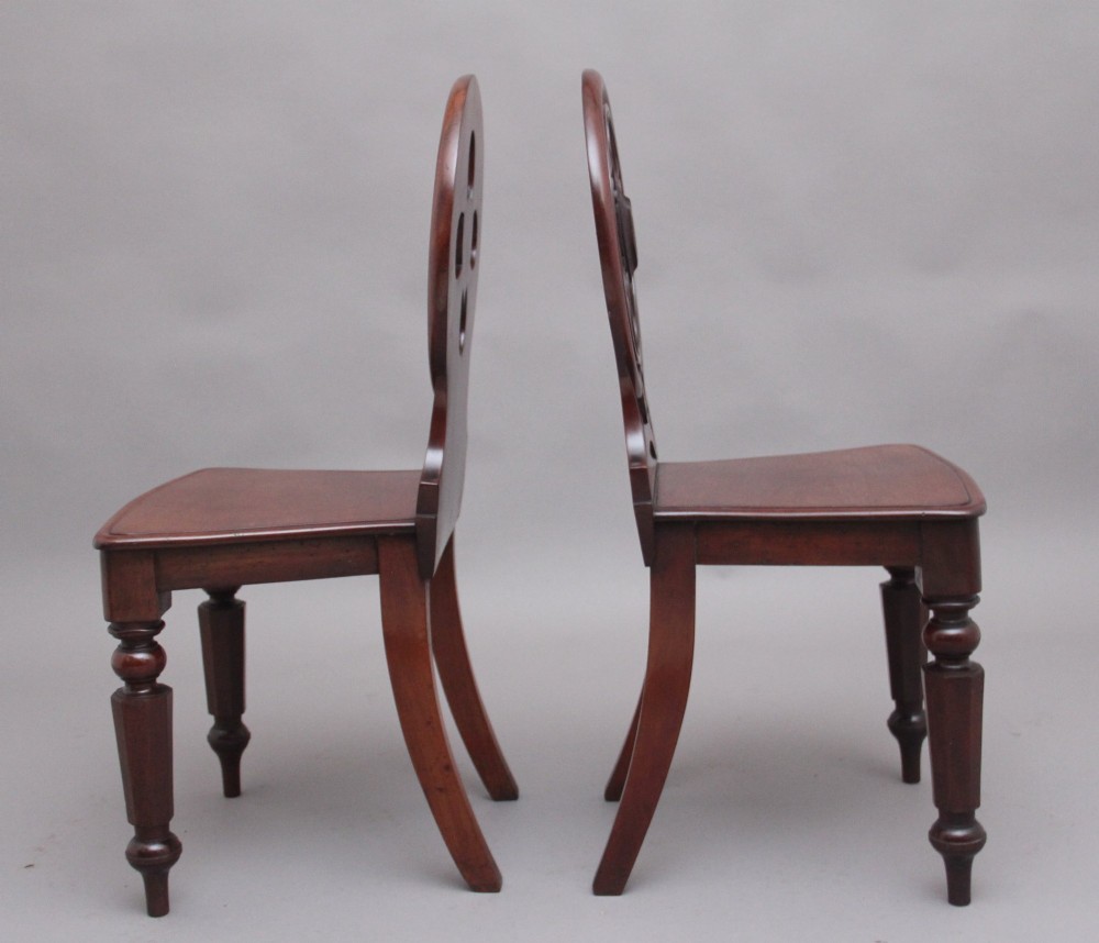 A pair of 19th-century mahogany hall chairs featuring circular carved backrests with Gothic tracery and central shield motifs above solid wooden seats and turned front legs. View V