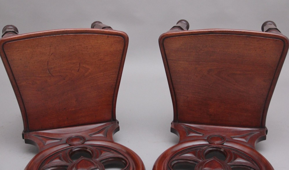 A pair of 19th-century mahogany hall chairs featuring circular carved backrests with Gothic tracery and central shield motifs above solid wooden seats and turned front legs. View VI