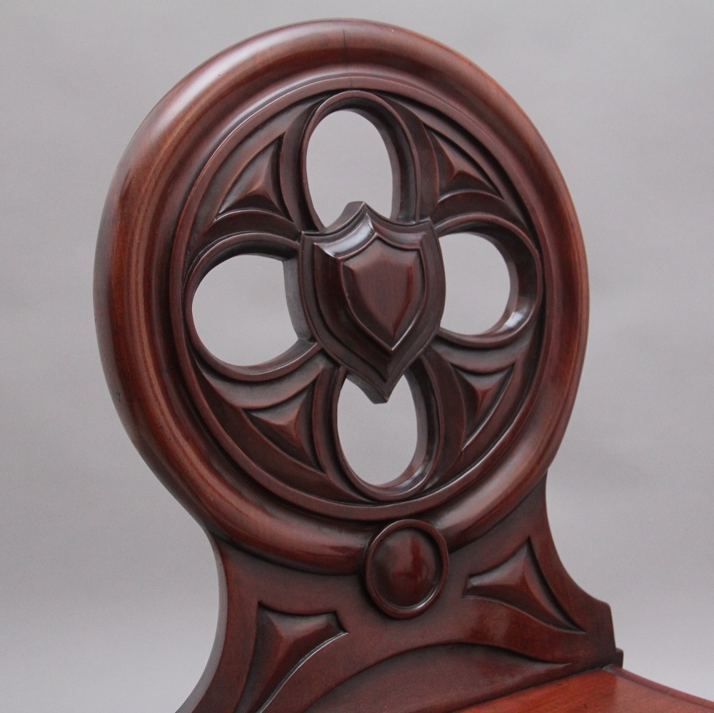 A pair of 19th-century mahogany hall chairs featuring circular carved backrests with Gothic tracery and central shield motifs above solid wooden seats and turned front legs. View VII