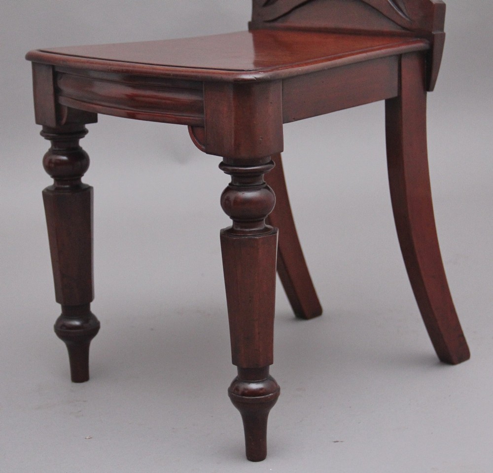 A pair of 19th-century mahogany hall chairs featuring circular carved backrests with Gothic tracery and central shield motifs above solid wooden seats and turned front legs. View VIII