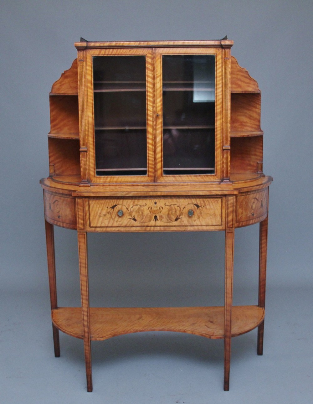 A 19th-century satinwood display cabinet featuring central glazed double doors, curved side shelves, a marquetry-inlaid drawer, and slender tapered legs with a lower undertier.