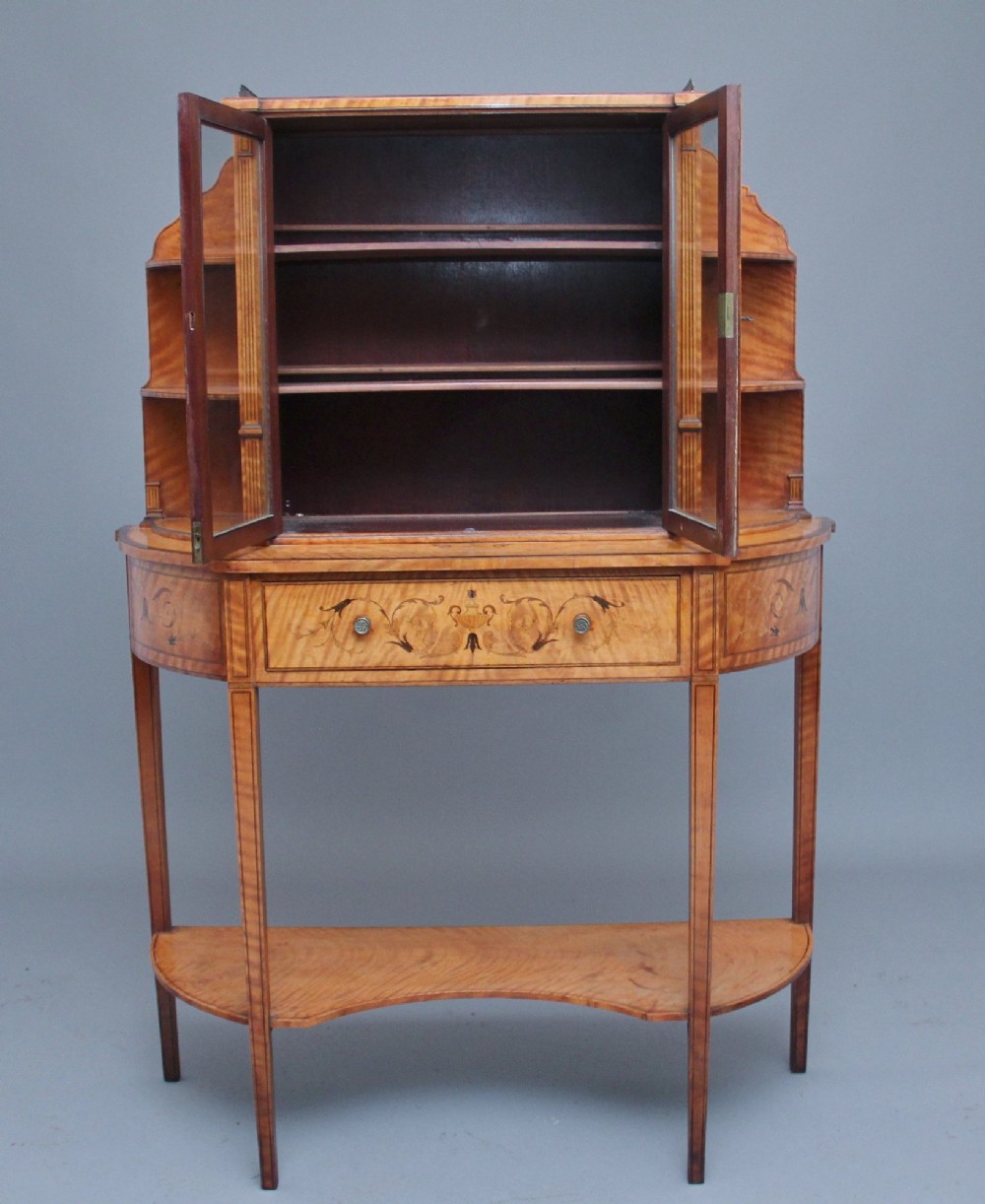 A 19th-century satinwood display cabinet featuring central glazed double doors, curved side shelves, a marquetry-inlaid drawer, and slender tapered legs with a lower undertier. View II