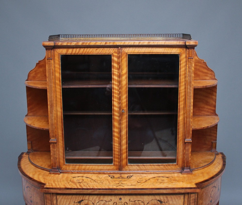 A 19th-century satinwood display cabinet featuring central glazed double doors, curved side shelves, a marquetry-inlaid drawer, and slender tapered legs with a lower undertier. View III