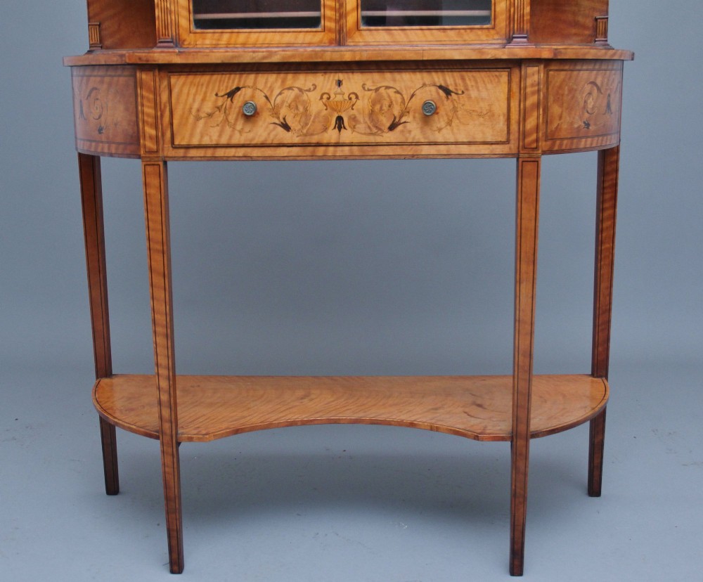 A 19th-century satinwood display cabinet featuring central glazed double doors, curved side shelves, a marquetry-inlaid drawer, and slender tapered legs with a lower undertier. View IV