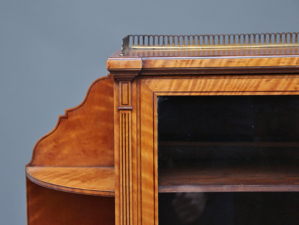 A 19th-century satinwood display cabinet featuring central glazed double doors, curved side shelves, a marquetry-inlaid drawer, and slender tapered legs with a lower undertier. View V