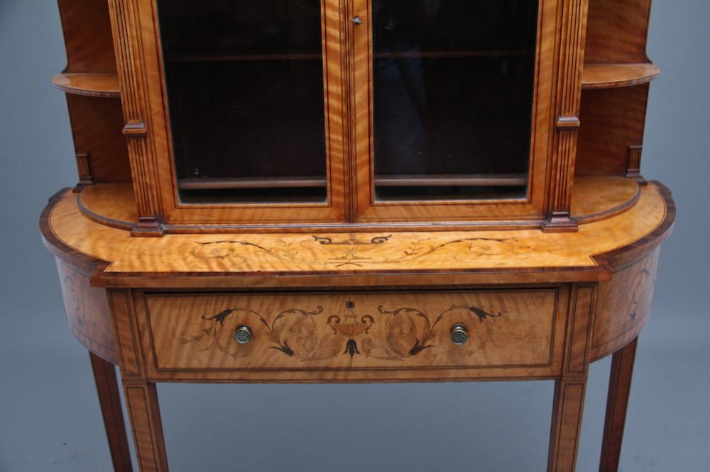 A 19th-century satinwood display cabinet featuring central glazed double doors, curved side shelves, a marquetry-inlaid drawer, and slender tapered legs with a lower undertier. View VI