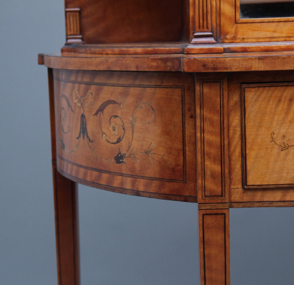 A 19th-century satinwood display cabinet featuring central glazed double doors, curved side shelves, a marquetry-inlaid drawer, and slender tapered legs with a lower undertier. View VII